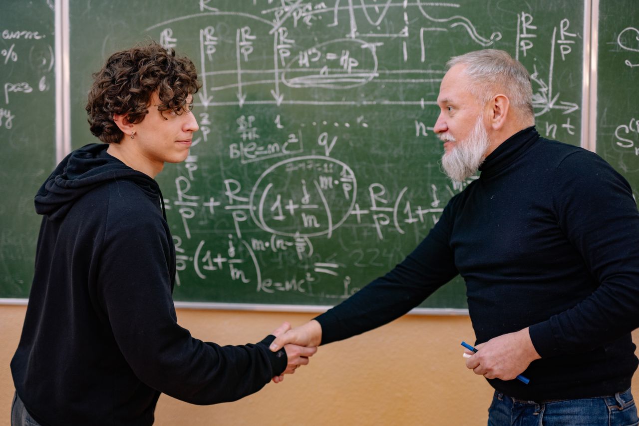 A professor and student shake hands in front of a chalkboard filled with mathematical formulas, symbolizing education and mentorship.